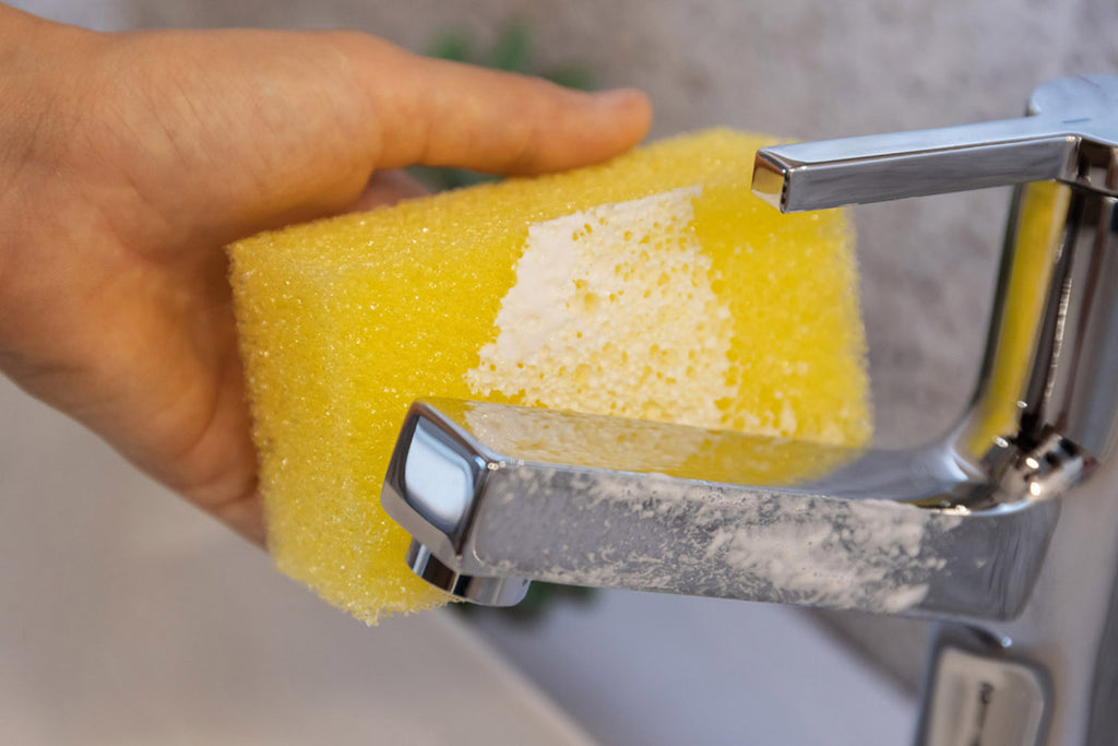 spugna gialla in mano che pulisce il rubinetto del bagno con pasta bianco del detergente